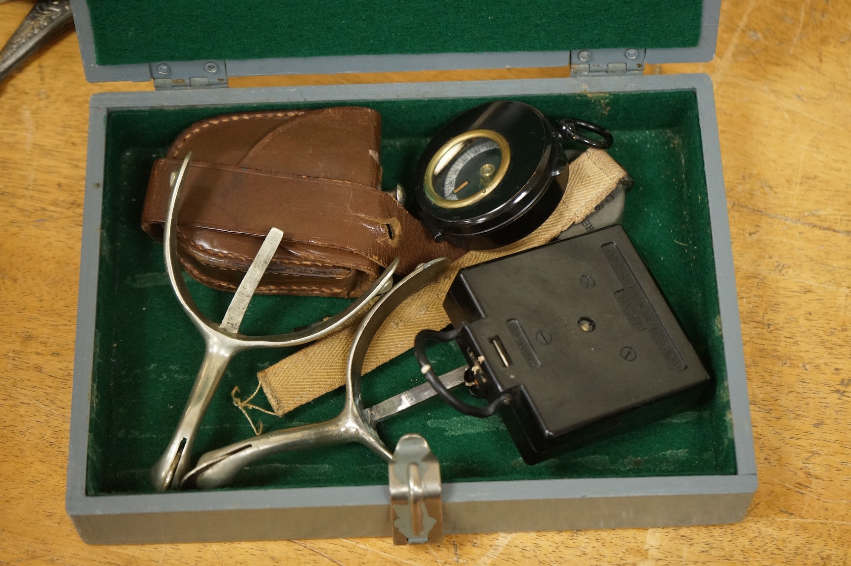 Three WWII military compasses including a Magnetic Marching Mark One compass, a small compass on a wrist strap and another in a leather case, together with a pair of steel spurs and a small grey painted wooden military b